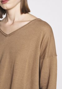 Jeune personne portant un pull à col en V ample de couleur marron clair, avec une couture d'épaule droite et des cheveux mi-longs et raides.