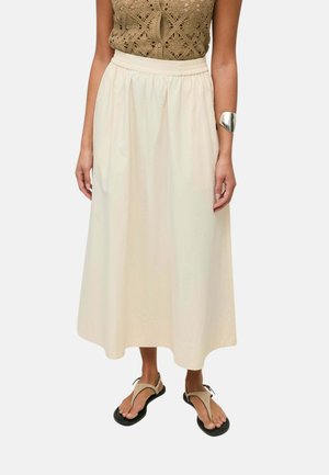 Mujer con top sin mangas de crochet color beige, falda maxi de cintura alta color crema, pulsera ancha plateada y sandalias beige con suelas negras.