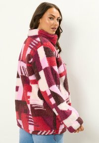 Fleecejacka med färgblockdesign i rosa, röd och brun nyanser, med hög krage och textil, mjukt material.