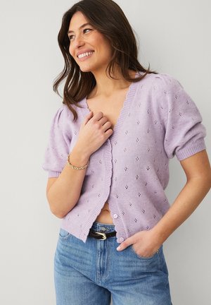 Kobieta uśmiechająca się, ubrana w jasnofioletowy krótki kardigan z dzianiny z małymi ażurowymi wzorami, w połączeniu z niebieskimi dżinsami i złotą bransoletką.