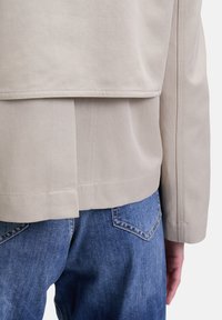 Beige canvas jack met een gelaagd ontwerp aan de achterkant, met zichtbare stiknaden en een ontspannen pasvorm, gecombineerd met blauwe jeans.