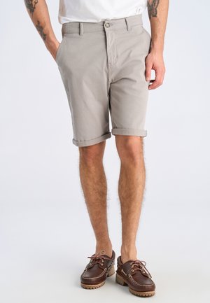 Hombre con pantalones cortos beige remangados, camisa blanca y zapatos náuticos de cuero marrón, de pie con una mano en el bolsillo contra un fondo liso.