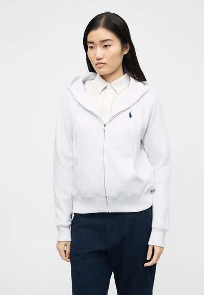 Νεαρή γυναίκα που φοράει λευκή ζακέτα με φερμουάρ πάνω από ένα ελαφρύ ριγέ πουκάμισο και σκούρο παντελόνι, στέκεται μπροστά σε απλό φόντο.