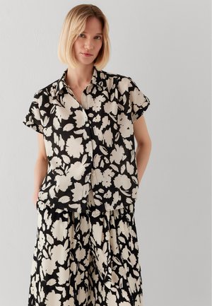 Woman with short blonde hair wearing a black and white floral short-sleeve button-up shirt and matching skirt against a plain background.
