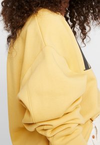 Persona con cabello rizado que lleva una sudadera amarilla suelta y pantalones blancos, vista de perfil con las mangas recogidas en las muñecas.