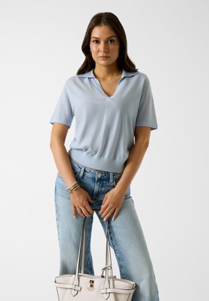 Frau trägt ein hellblaues, kurzärmeliges V-Ausschnitt-Oberteil und Jeans, hält eine weiße Lederhandtasche mit zwei Griffen vor einem einfarbigen Hintergrund.