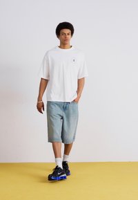 Camisa blanca de gran tamaño con un pequeño gráfico, combinada con pantalones cortos de denim azul claro. Zapatillas negras con acentos azules. Fondo simple.