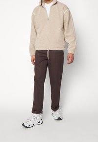 Beige fleecepullover med halvdämpning och sidodetaljer, parat med mörkbruna byxor och vita sneakers med svarta sulor. Avslappnad outfit.