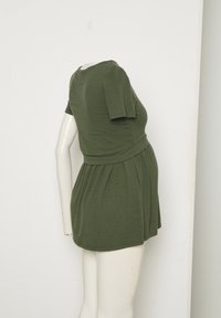 Mannequin wearing short-sleeve olive green dress with gathered waist, standing against plain white wall, headless and armless on one side.