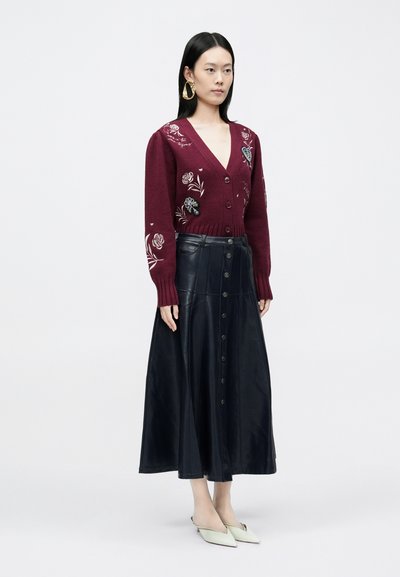 Bordó kötött kardigán virágos hímzéssel, fekete bőr középhosszú szoknyával, amely gombokkal díszített, és világoszöld hegyes orrú balerinacipőkkel párosítva.