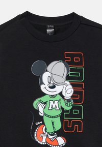 Mickey Mouse in a green outfit with an "M" logo, wearing a cap, poses playfully. The shirt is black with colorful "Adidas" text.