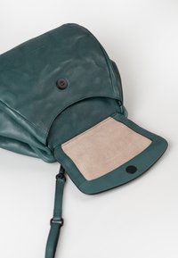 Zaino in pelle color teal con una fodera interna morbida di colore chiaro, dotato di una chiusura a scatto nera. Il design include una tracolla removibile.