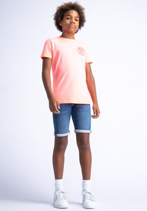 Camiseta rosa claro con logo redondo, combinada con shorts de denim con borde doblado. Zapatillas blancas con cordones y calcetines blancos hasta el tobillo.