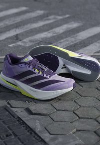Ζευγάρι μωβ παπούτσια τρεξίματος Adidas Adizero με σκουρόχρωμες ρίγες και νέον κίτρινη λεπτομέρεια σε πλακόστρωτο δρόμο.