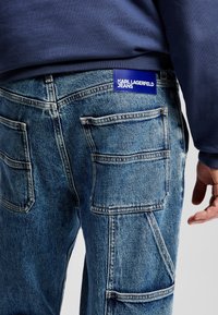 Calças de ganga azul com múltimos bolsos, costura visível e um rótulo azul com a inscrição "KARL LAGERFELD JEANS" perto da cintura.