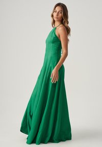 Abito lungo verde senza maniche con corpino aderente e gonna fluente. Realizzato in tessuto morbido con drappeggio rilassato e design minimalista.