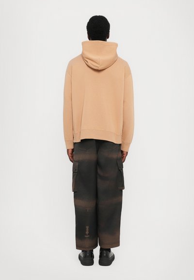 Beige huvtröja med sänkta axlar, tillsammans med svarta cargobyxor med stora sidofickor och en urbleknad textur. Svarta skor fulländar looken.