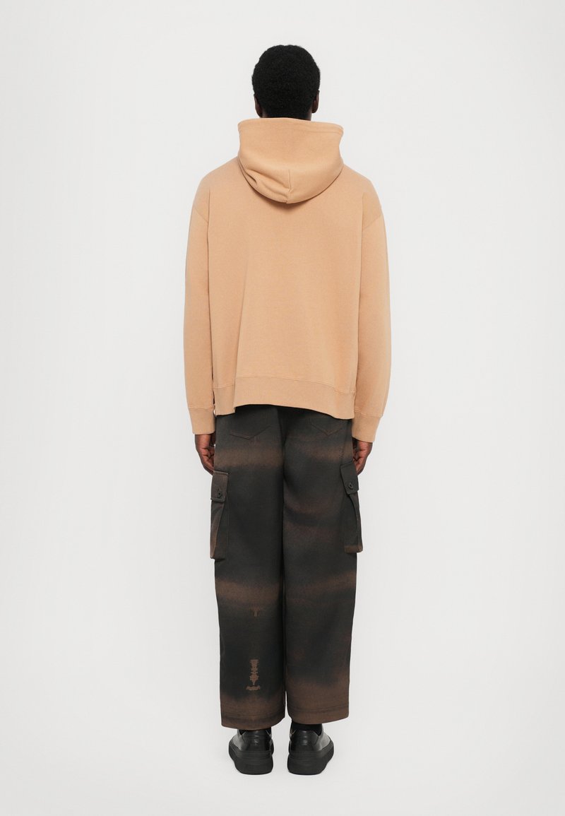 Beige Kapuzenpullover mit überschnittenen Schultern, kombiniert mit schwarzer Cargo-Hose mit großen Seitentaschen und verwaschener Textur. Schwarze Schuhe runden den Look ab.