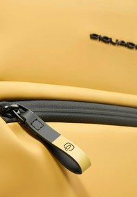 Detalle de una mochila sintética amarilla con una cremallera gris y una lengüeta de tirador negra. Presenta una textura suave y un diseño minimalista.