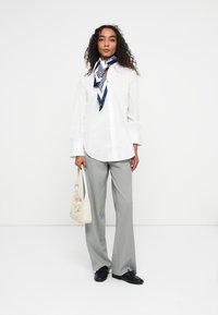 Biała koszula na guziki z subtelną teksturą, szare spodnie w paski, granatowo-biała wzorzysta chusta oraz jasnobeżowa torba z detalem w postaci kokardy.