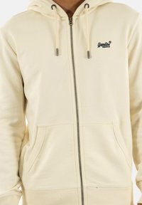 Creme zip-up hoodie med foret hætte, dobbelte lommer og ribbede ærmer. Har et lille logo i mørkeblåt på venstre bryst. Blød tekstur.