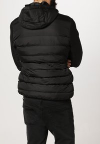 Chaqueta acolchada negra con capucha, que presenta secciones horizontales acolchadas y una textura suave. Llevada sobre jeans oscuros, vista desde la parte de atrás.