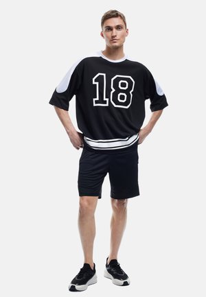 Canotta sportiva nera con accenti bianchi e il numero 18 sul davanti, abbinata a pantaloni corti neri e sneakers nere.