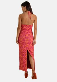 Vestido maxi en tela rosa y naranja con un diseño de cuello halter, que presenta una abertura lateral y patrones intrincados. Textura suave. Tacones marrones.
