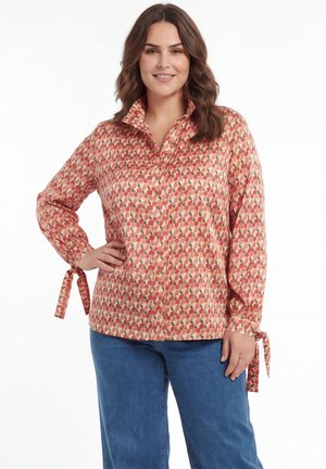 Vrouw draagt een bloemenblouse met lange mouwen en strikmanchetten, gecombineerd met een blauwe spijkerbroek, staand met één hand in de zij en glimlachend.