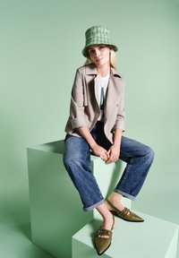 Beige doppeltknöpfige Jacke über einem weißen T-Shirt, blauen Jeans, grünem Tweed-Bucket-Hat und olivgrünen Ballerinas mit Nietenakzenten.