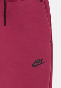Nike tepláky v hlbokej ružovej farbe s jemnou textúrou. Obsahujú čierne logo a šnúrku s čiernymi koncami. Úzky, zúžený strih.