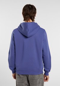 Persona vista da dietro che indossa un felpa blu e pantaloni grigi su uno sfondo chiaro e semplice.