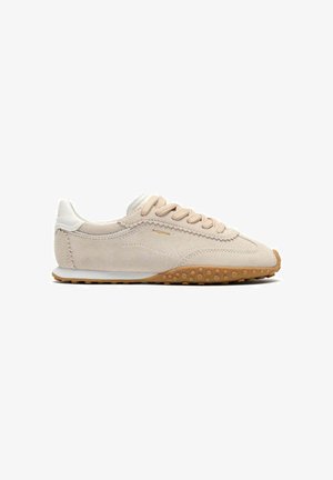 Zapatilla baja de ante beige con lengüeta blanca en el talón, cordones beige y suela de goma marrón texturizada, vista lateral sobre fondo blanco.
