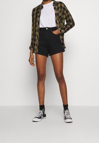 Persona vestida con pantalones cortos negros enrollados, camiseta blanca, una camisa a cuadros en tonos oliva y negro, calcetas negras y zapatillas altas negras, de pie contra un fondo blanco.