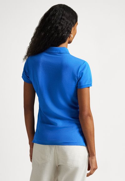 Μπλε πόλο μπλούζα με γιακά, κοντά μανίκια, υφασμένο ύφασμα και εφαρμοστή γραμμή. Φορεμένη με ανοιχτόχρωμο παντελόνι, σε προβολή από πίσω.