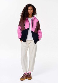 Kolorowy hoodie z zamkiem błyskawicznym w różowym, brązowym i czarnym kolorze, z kieszeniami z przodu i sznurkiem do regulacji. W zestawie z białą koszulą i kremowymi spodniami. Brązowe sneakersy.