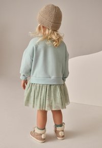 Pulover bleu deschis cu mâneci tip balon, purtat peste o fustă plisată verde mentă cu modele florale mici. Accesoriile includ o căciulă tricotată bej și cizme maro cu blănuri sintetice și pompoane.