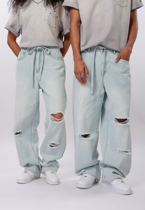 Dvaja ľudia stojaci vedľa seba, oblečení v svetlomodrých roztrhaných voľných džínsoch so sťahovacím pásom v páse, sivých tričkách s vyhrnutými rukávmi a bielych teniskách.