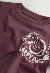 T-shirt en coton bordeaux avec impression graphique blanche présentant des personnages souriants, une spirale et le texte "JOURS HEUREUX" avec des accents. Design à col rond.