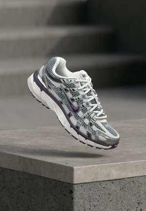 Biely a sivý Nike teniska s fialovými akcentmi, vznášajúci sa nad betónovou rímsou s rozmazanými schodmi v pozadí.