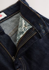 Jeans in denim blu scuro con zip aperta che rivela una fodera interna decorata e cuciture marroni visibili intorno alla vita e alla asola.