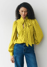 Gelbe Bluse mit strukturierten Polka-Dots, einem tiefen V-Ausschnitt und einer Schleife am Kragen. Verfügt über voluminöse Puffärmel. Getragen mit blauen Jeans.