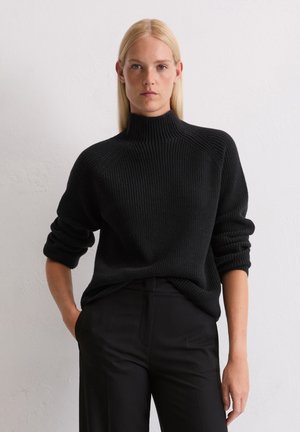 Svart ribbad stickad tröja med hög halsringning och uppvikta ärmar, tillsammans med mörka byxor. Tyget verkar tjockt och texturerat.