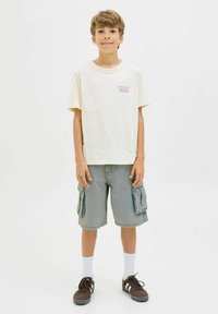 T-shirt beige clair à manches courtes avec logo rouge, associé à un short cargo gris ample avec poches latérales, des chaussettes blanches et des baskets noires.