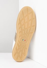 Semelle de chaussure avec une surface texturée et en caoutchouc beige, présentant un emblème circulaire. La partie supérieure en blanc avec une bande en argent métallique.
