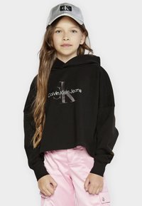 Felpa nera corta con logo di Calvin Klein Jeans in argento. Abbinata a pantaloni di satin rosa chiaro e un cappellino grigio. Capelli lunghi e ondulati.