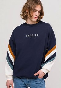 Felpa blu navy con vestibilità oversize, maniche a righe a contrasto in arancione bruciato, bianco e teal. Presenta il logo "KAOTIKO" sul davanti.