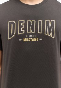 Tamsiai pilka marškinėliai su trumpomis rankovėmis. Ant jų yra ryškus "DENIM" tekstas baltos ir geltonos spalvos, o po juo yra "CLASSICS" ir prekės ženklas "MUSTANG".