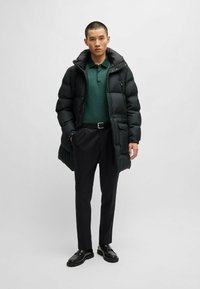 Black puffer jacket with a hood and front pockets, worn over a dark green polo shirt, paired with tailored black trousers and shiny black shoes.