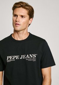 Melnas kokvilnas T-krekls ar īsām piedurknēm, kuram ir balts drukāts zīmols "PEPE JEANS PORTOBELLO RD" un smalks auduma raksts.